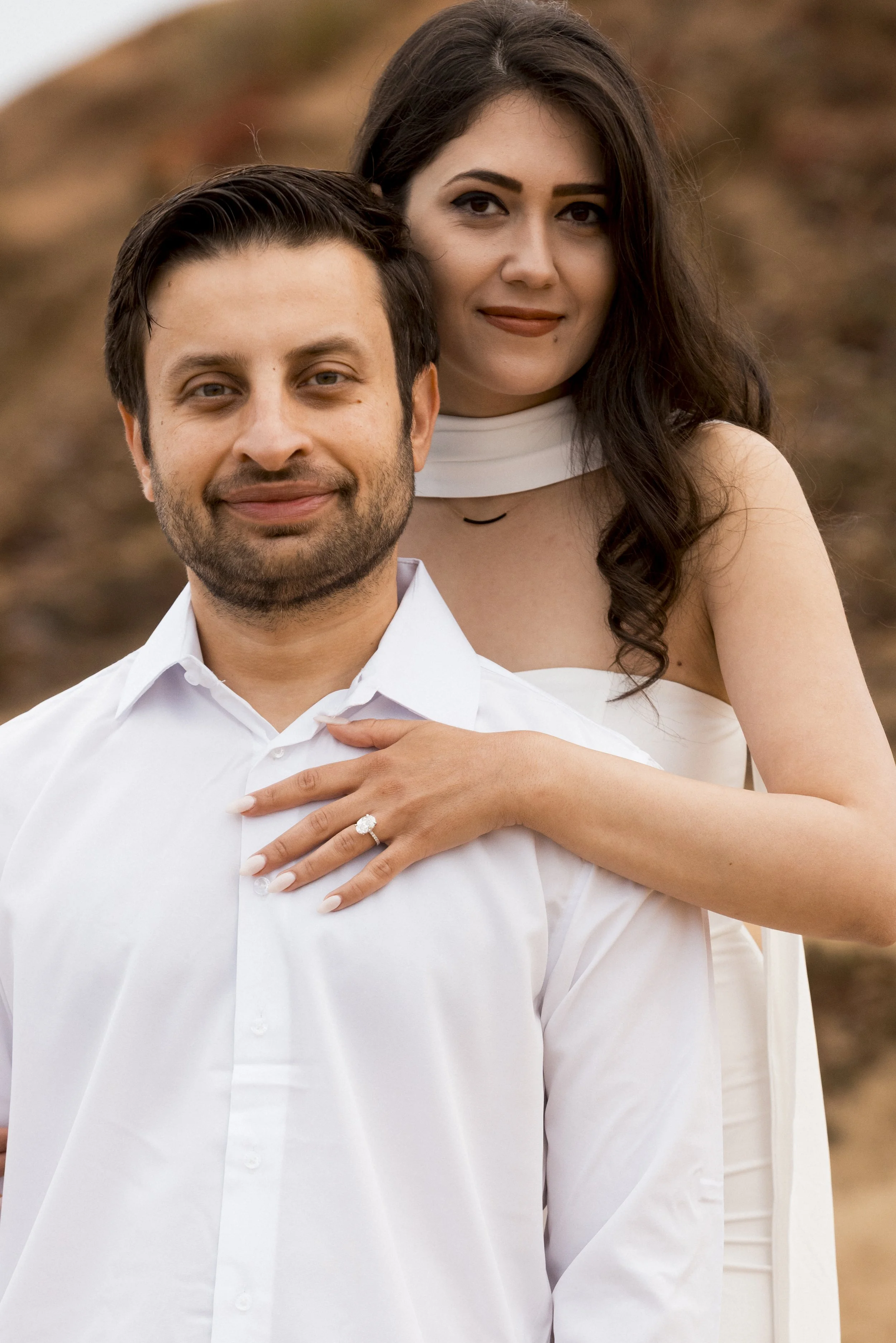 A couple looking into each other's eyes and smiling outdoors, with a church tower and trees in the background.
