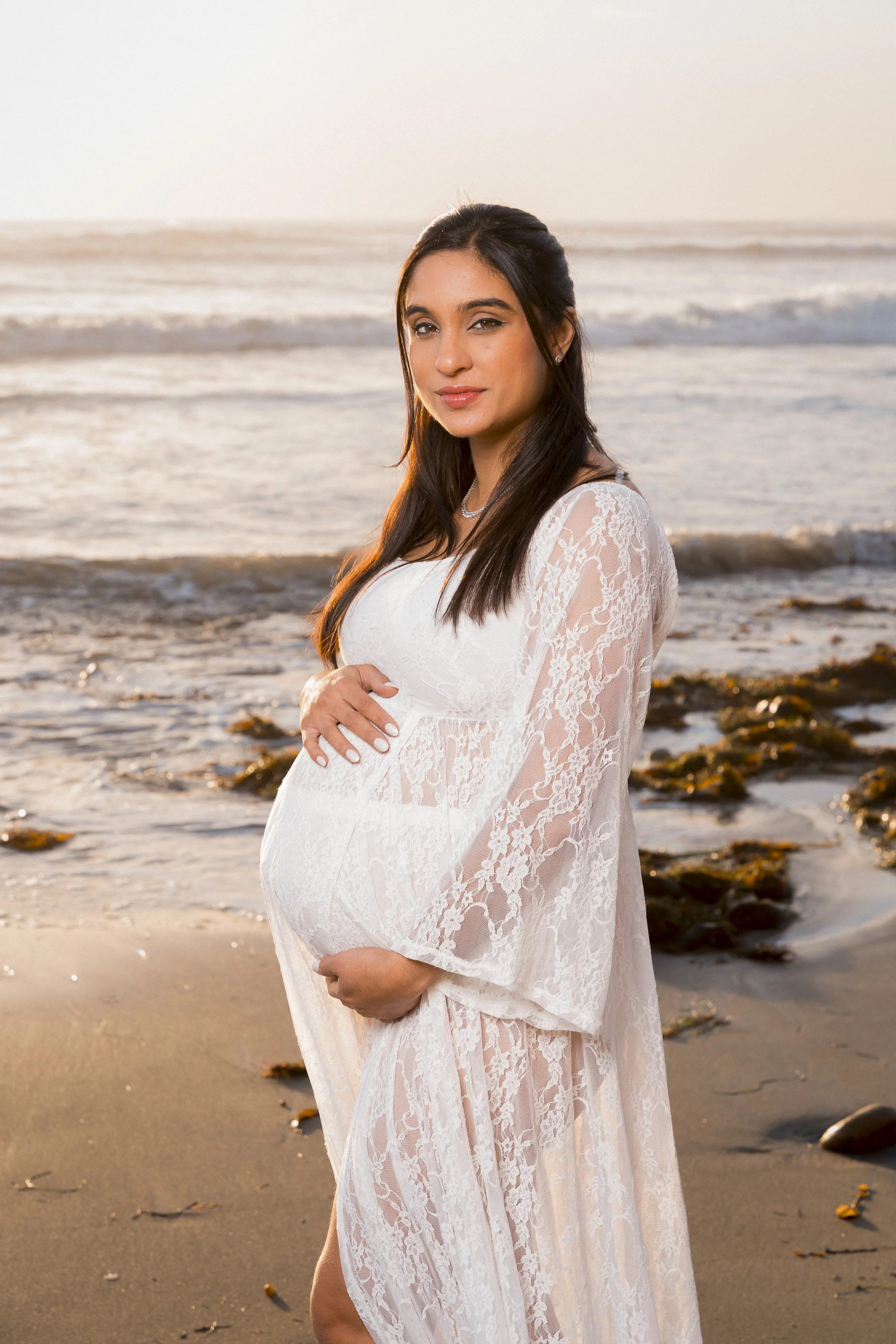 A pregnant woman in a white lace dress standing on a sandy beach at sunset, holding her baby bump and looking at the camera.