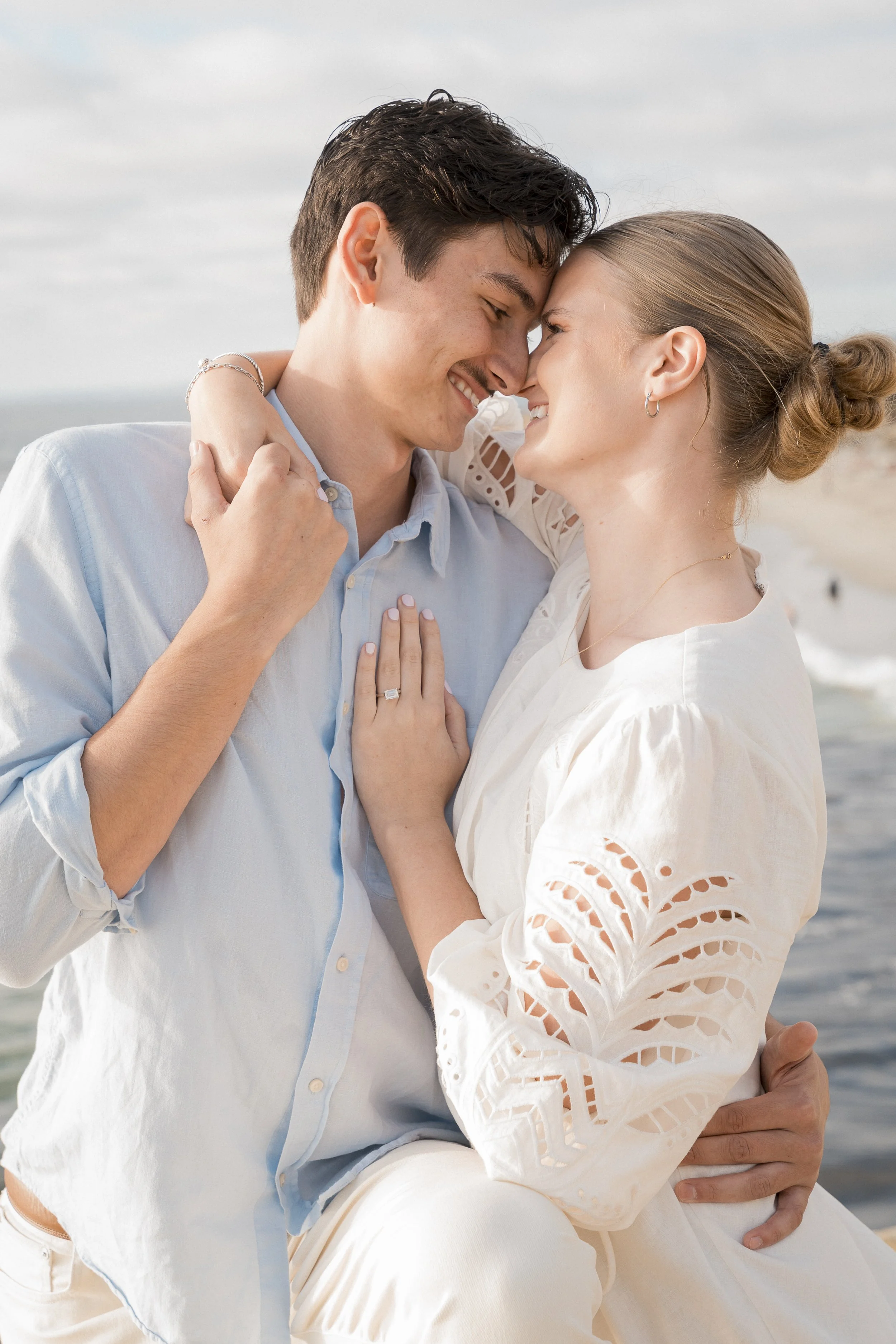 A happy couple sharing an intimate moment on the beach, smiling and touching foreheads, with ocean waves in the background.