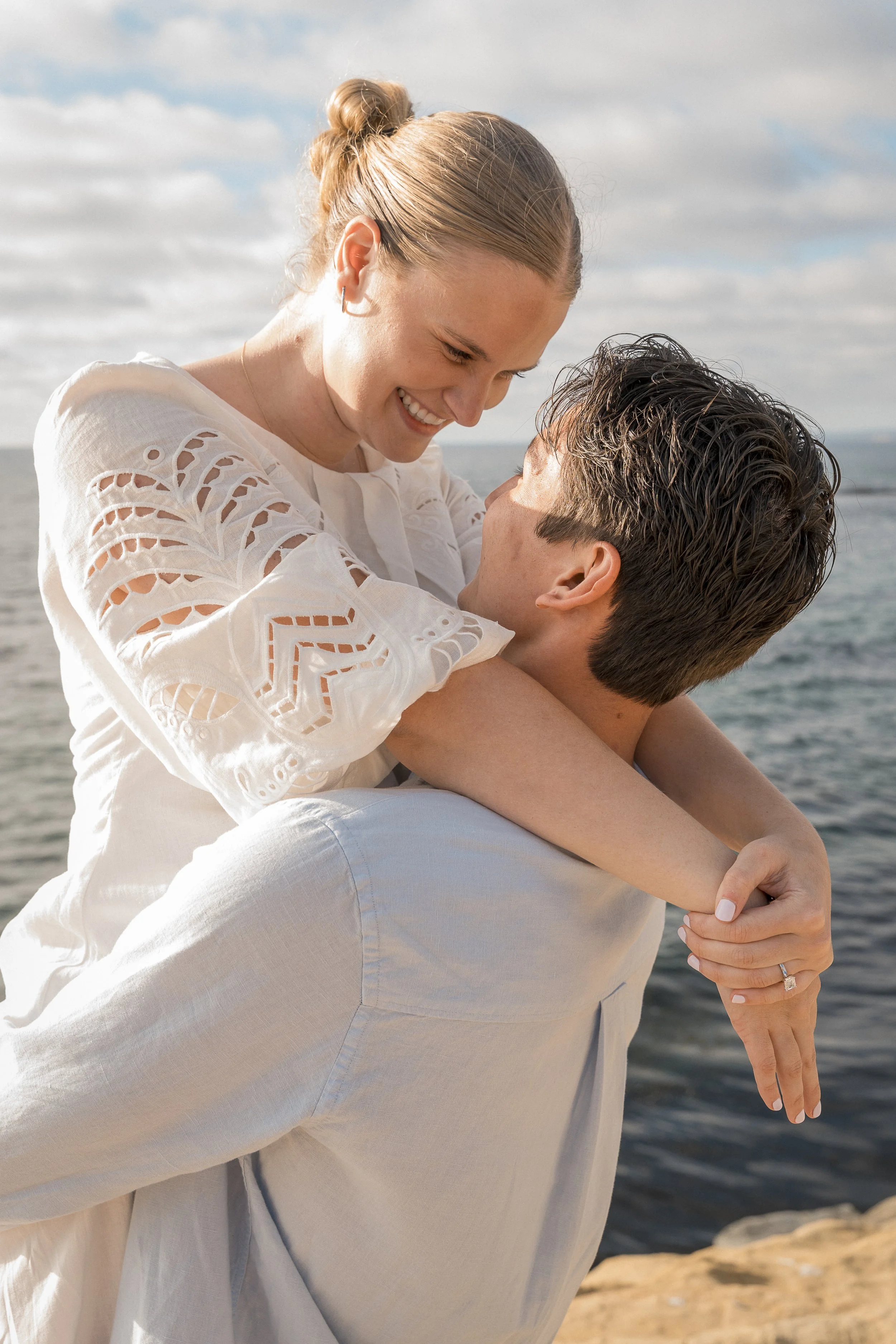 A smiling woman with long brown hair on a man’s back at the beach, with a rocky cliff in the background.