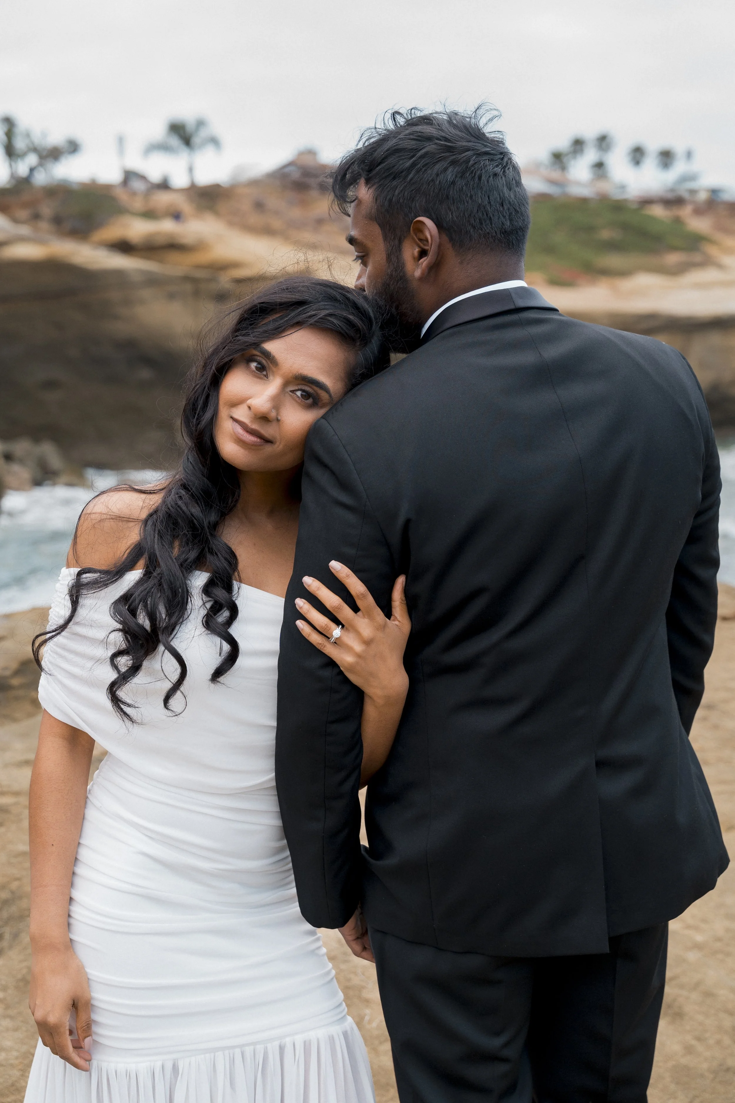 A happy couple looks into each other's eyes in an outdoor park with a building tower in the background.
