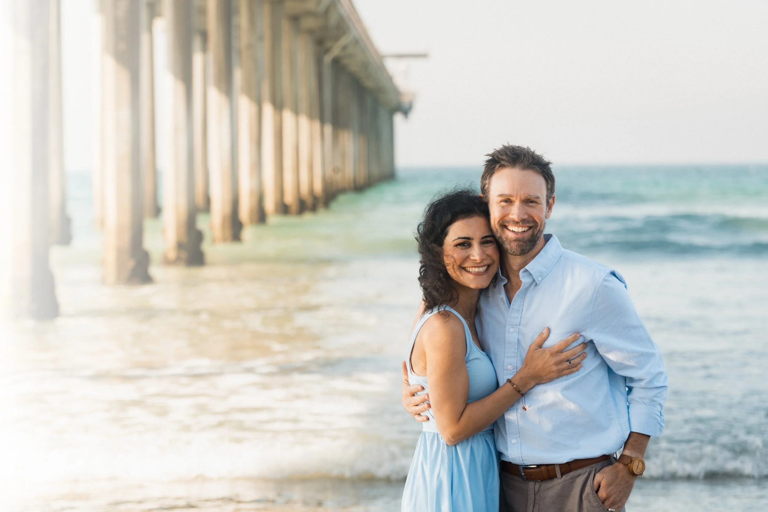 A couple dressed in formal attire standing on a beach at sunset, smiling and gazing into each other's eyes with a rainbow in the sky behind them.