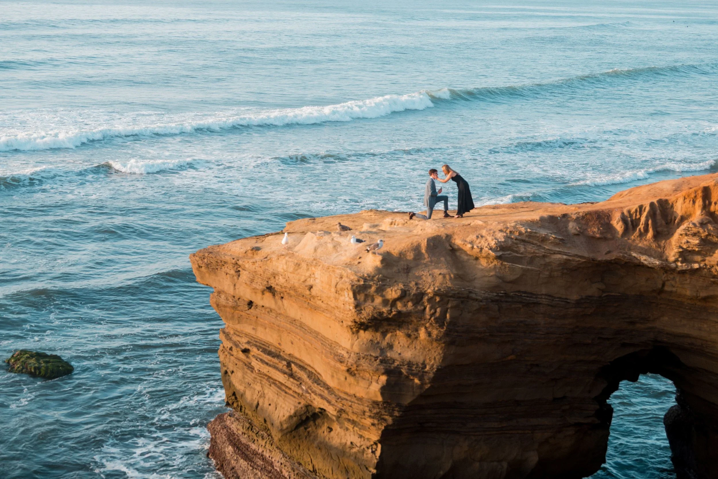 A couple on a rocky cliff by the ocean, with one person kneeling and proposing to the other. The ocean waves can be seen in the background.