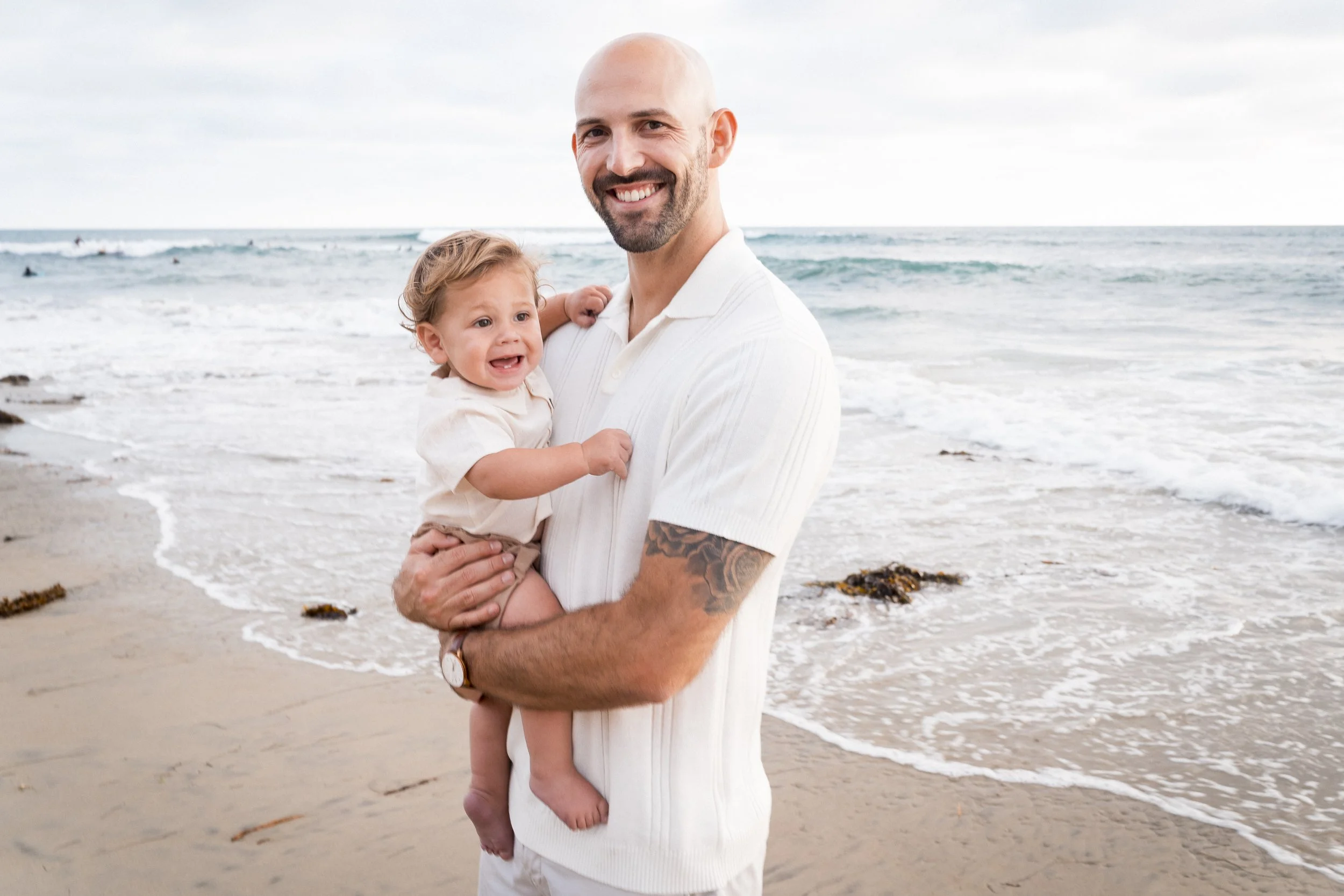 A man and a young boy walking barefoot on a sandy beach holding hands, with the ocean and blue sky in the background.