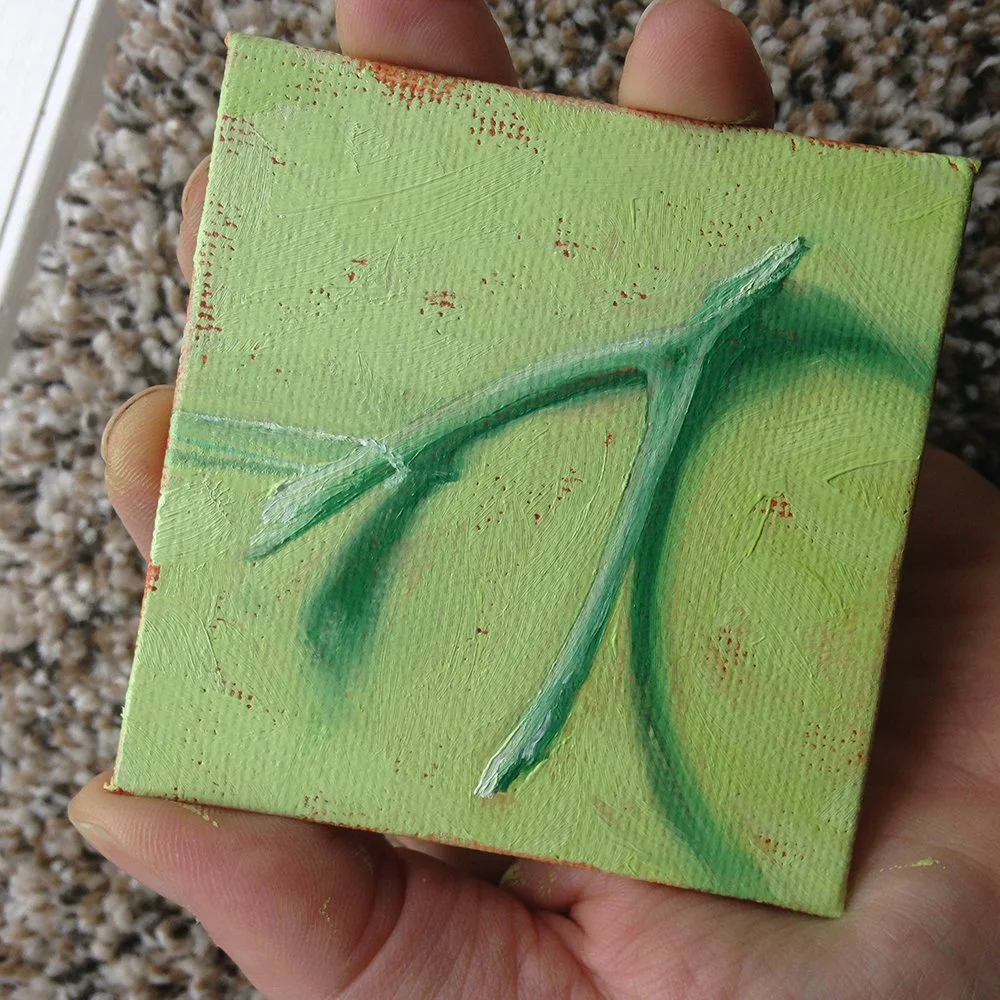 Close-up of a small painted canvas with a green monochromatic painting of a wishbone, held in someone's hand over a beige carpet.