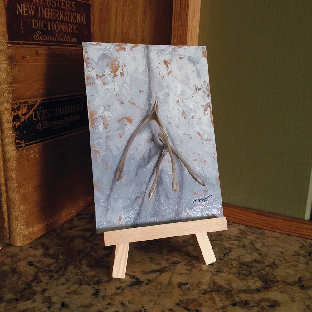 A small painting on a wooden easel depicts a minimalist, textured depiction of two wishbones on a gray and white abstract background. The easel is placed on a granite countertop, with a shelf of books behind it.