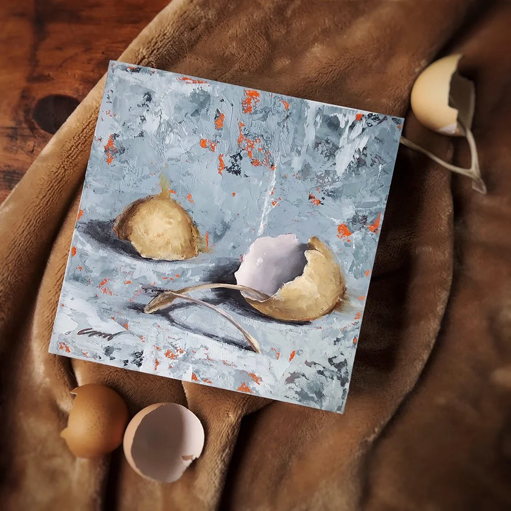 A small, square, painted panel depicting two eggshells and a wishbone on a textured surface. The artwork is placed on a soft, brown fabric, surrounded by cracked eggshells and a wishbone.
