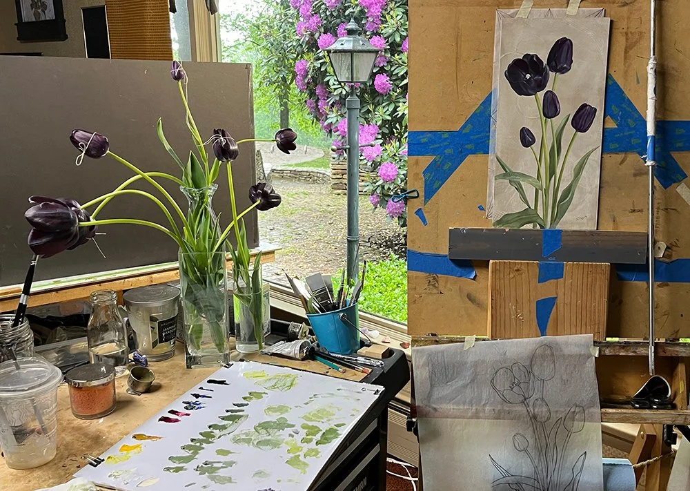 Black tulips bending and following the light on the artist's work station, next to the actual oil painting of them.