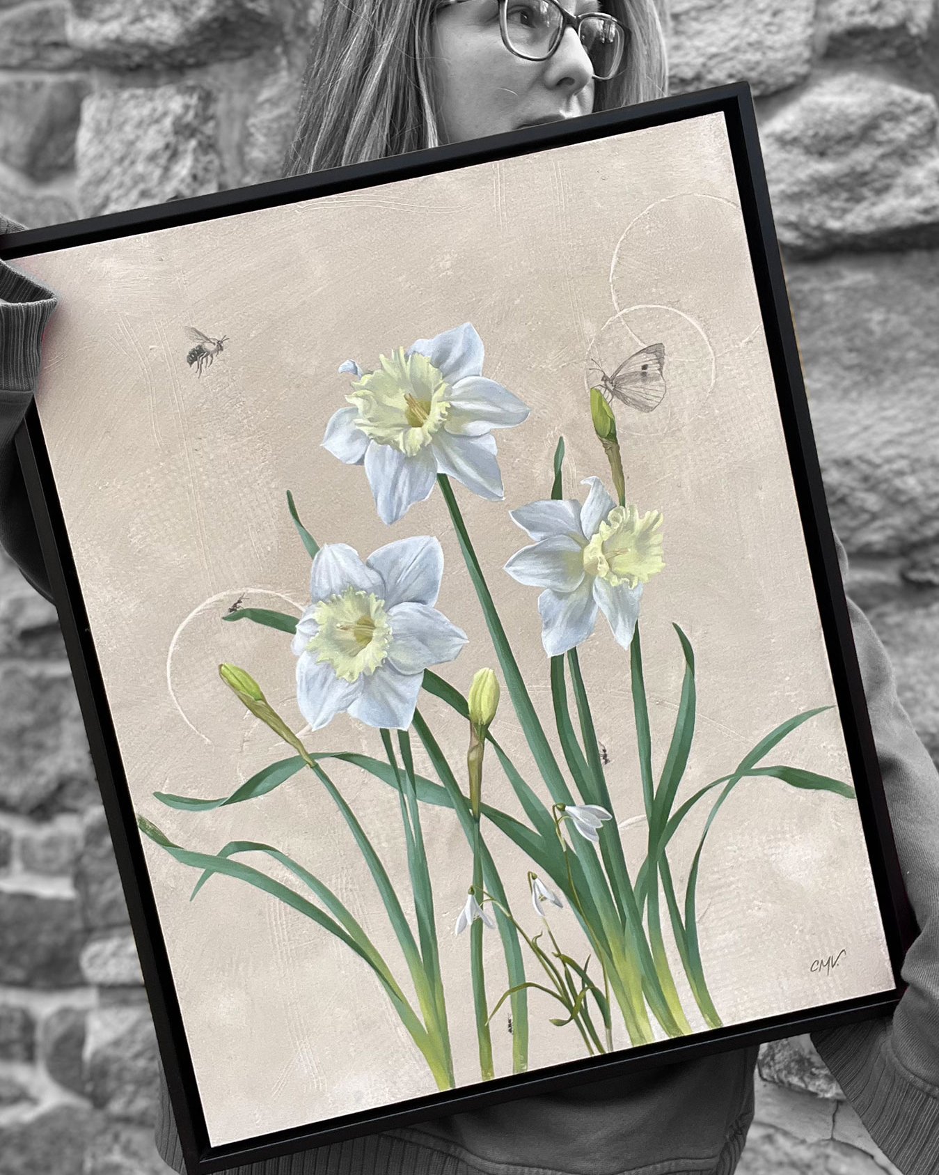 A painting of white and pale yellow day lilies and three snowdrop flowers with ants, a bee, and a butterfly framed in black and held by Christine Mercer-Vernon.