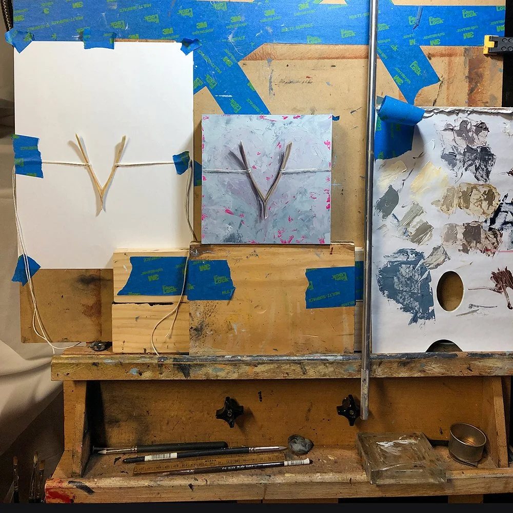 Art studio workspace with a side by side comparison of a wishbone and the painting of it, taped to a wooden work surface with various art supplies, including brushes and paints, and an overall cluttered environment.
