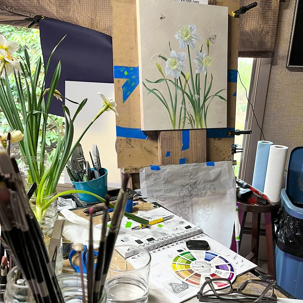 The artist's studio with a work in progress daffodil painting on the easel and a vase of daffodils far left.