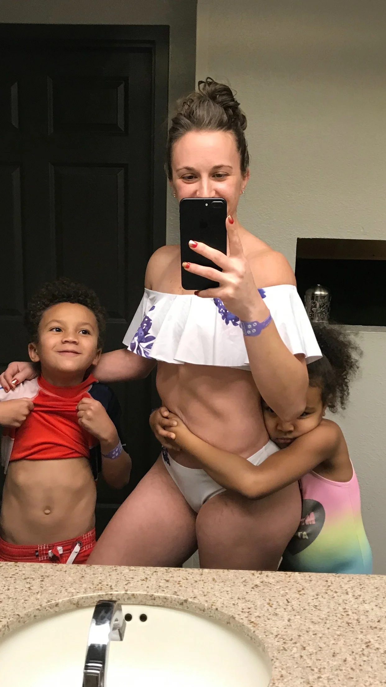 A woman taking a mirror selfie in a bathroom with two children hugging her on either side.