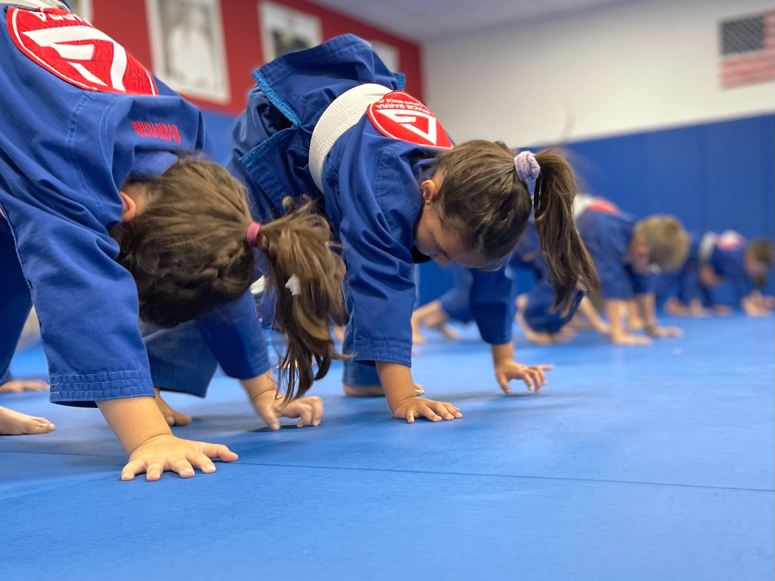 Children practicing jiu-jitsu in blue uniforms inside a dojo, performing a warm-up exercise on a blue mat.