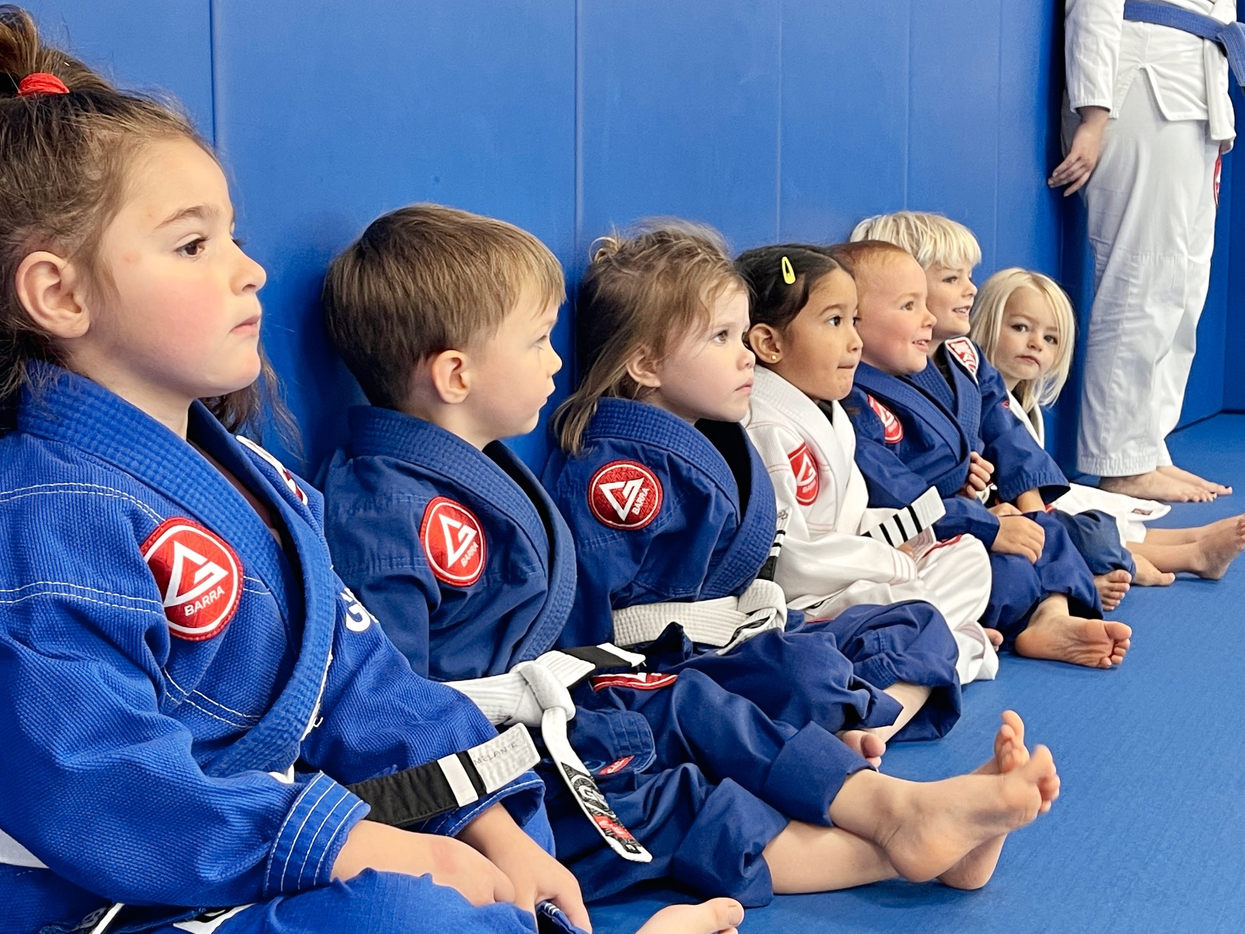A group of young children in blue and white martial arts uniforms sitting on a mat, with focused expressions, in a training setting.