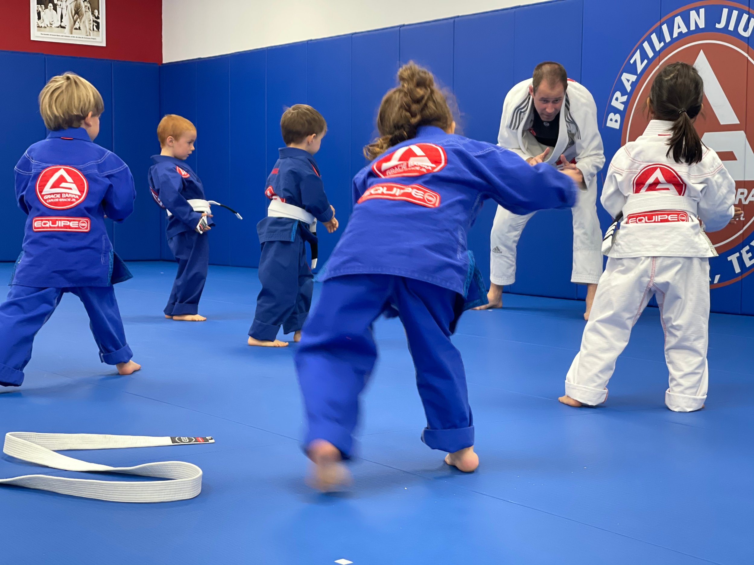 Children in blue and white jiu-jitsu uniforms practicing on a mat with an instructor in a martial arts studio.