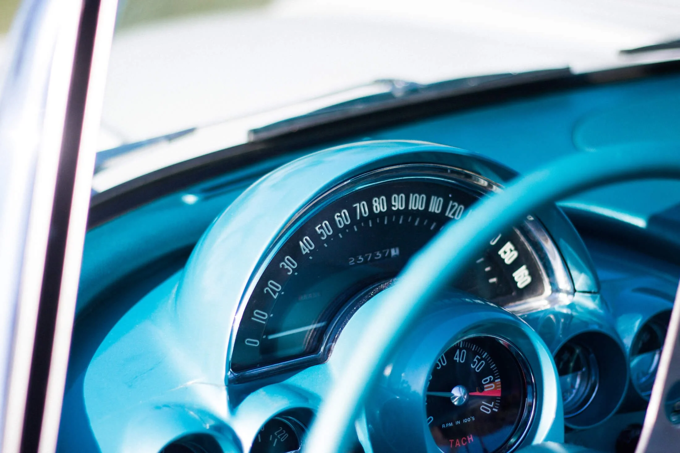 Close-up of a vintage white and turquoise 1959 Corvette convertible dashboard with a turquiose background, showing a speedometer, tachometer, and other instrument gauges.