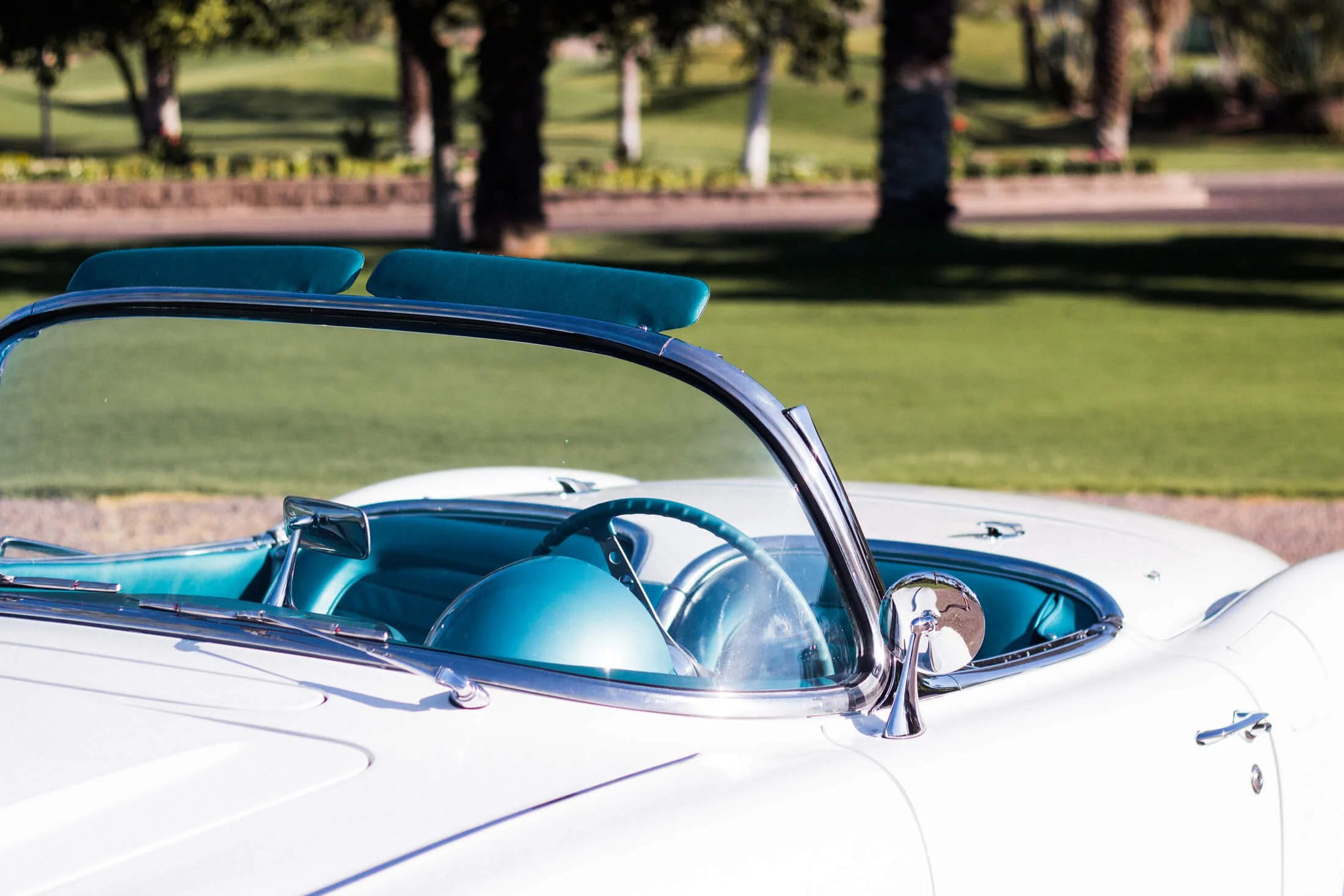 Close-up of a vintage white and turquoise 1959 Corvette convertible car with teal interior and teal headrests, parked on a grassy area with trees in the background.