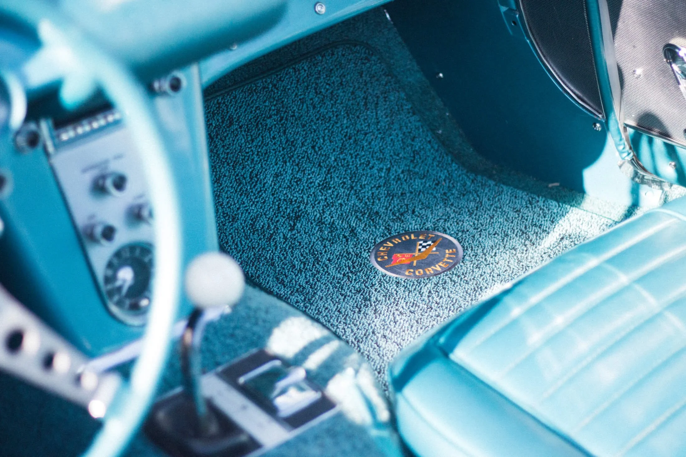 The interior of a vintage 1959 Chevrolet Corvette with a blue dashboard and matching blue seats. The floor mat features the Chevrolet Corvette logo with a checkered flag.