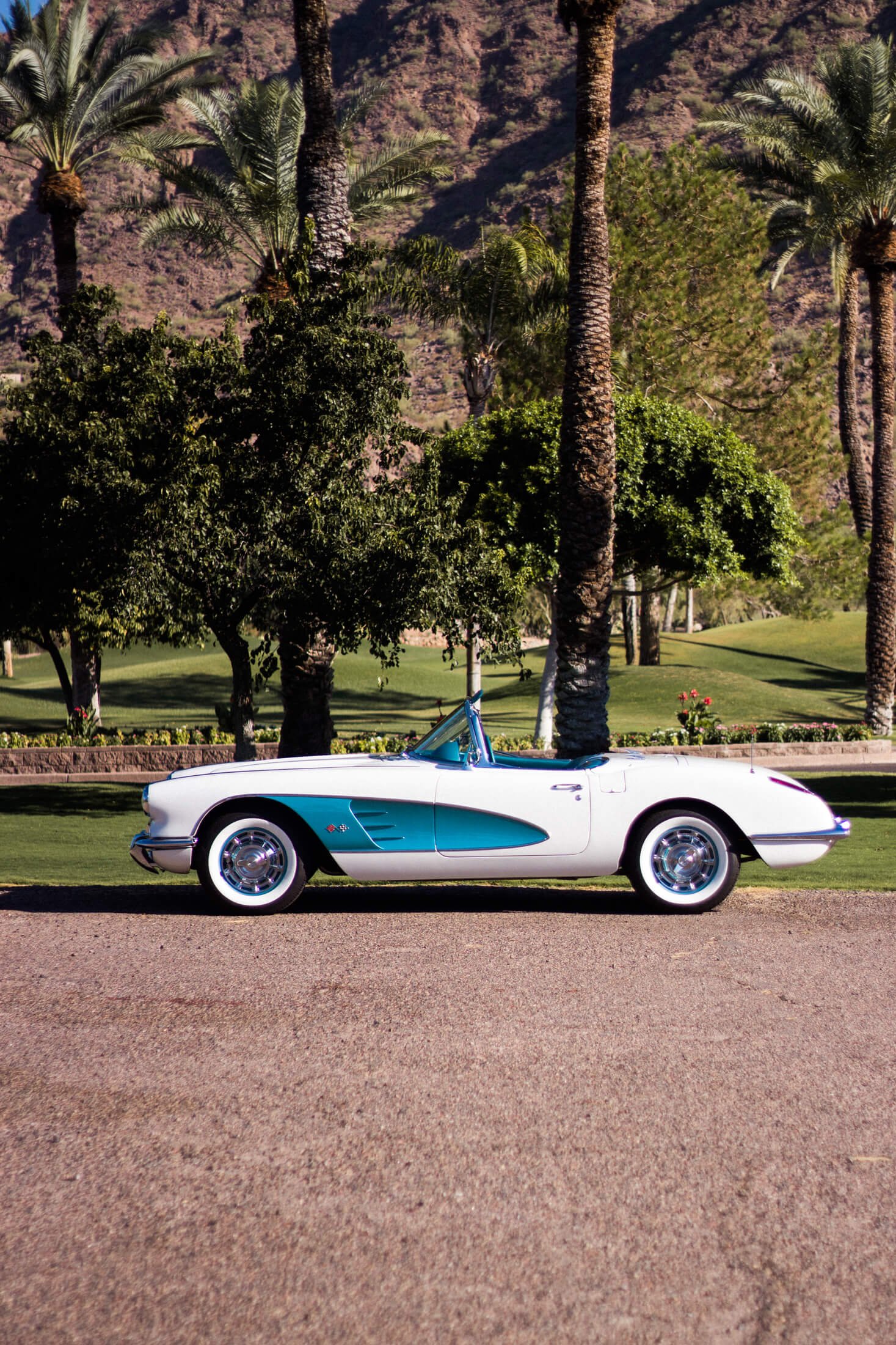 A vintage white and teal 1959 Chevrolet Corvette car parked on a gravel road with a lush green park, palm trees, and mountains in the background.