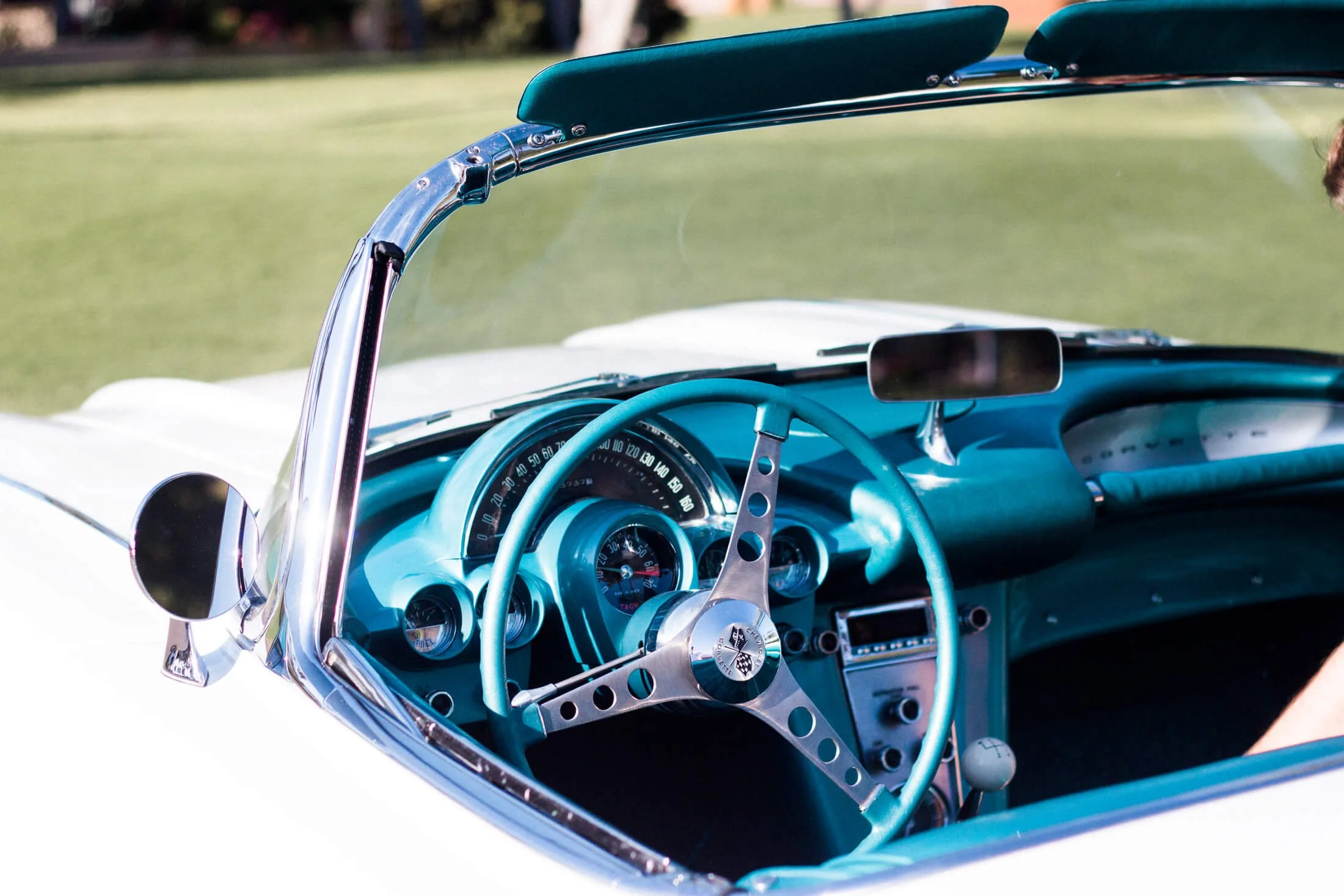 Close-up of the interior dashboard and steering wheel of a vintage white and turquoise 1959 Corvette convertible, with a view of the speedometer and side mirror.