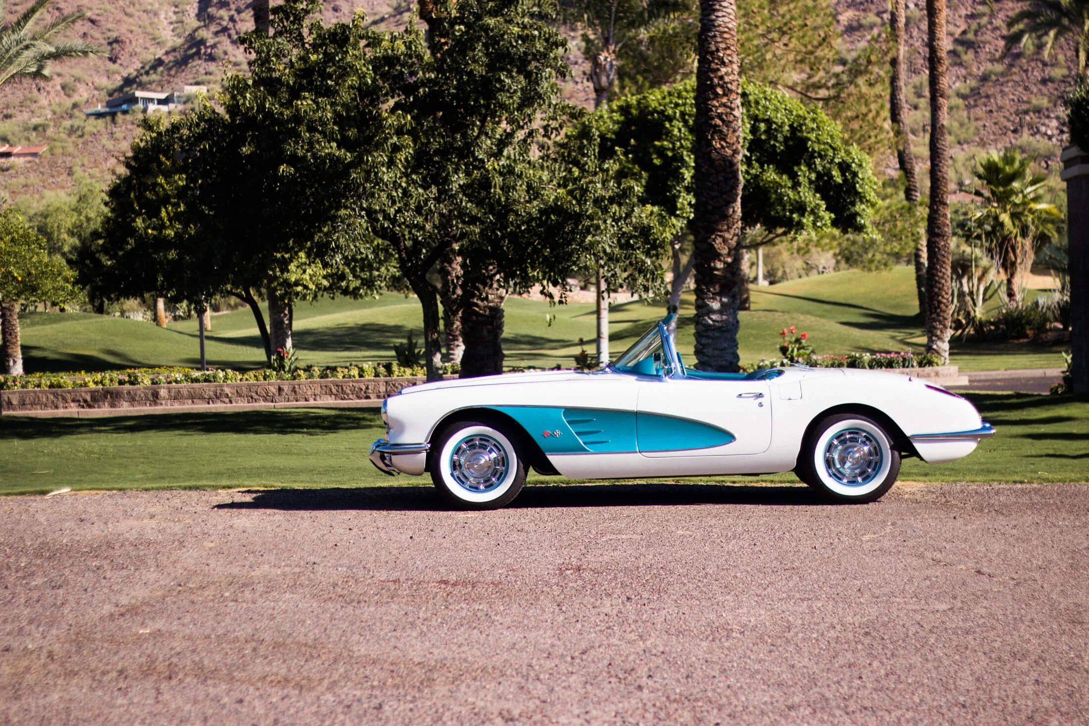 A vintage white and turquoise 1959 Chevrolet Corvette convertible with blue accents parked on a gravel surface, surrounded by lush green trees and a well-manicured lawn, with a hillside and houses in the background.