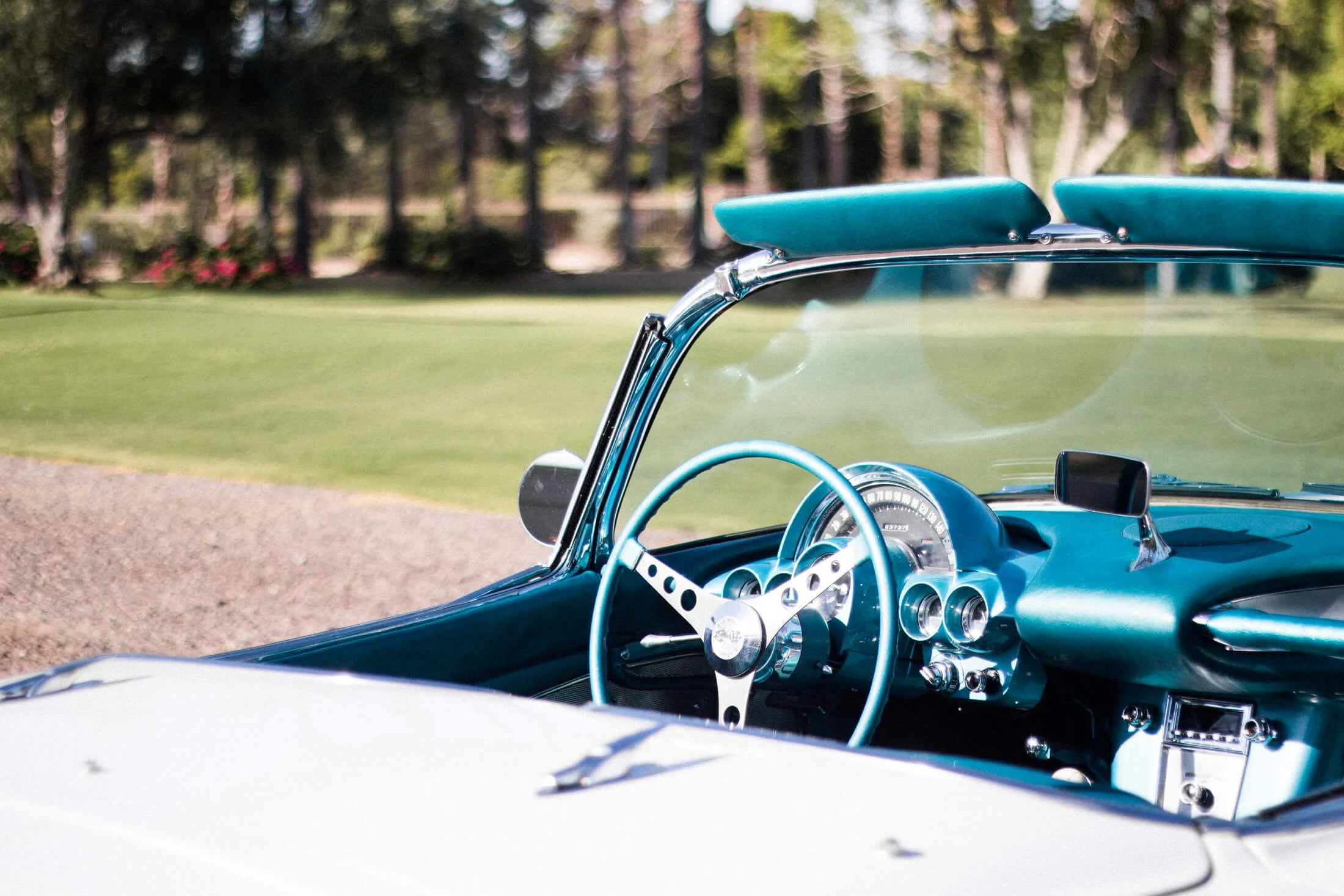 Close-up of a vintage white and turquoise 1959 Chevrolet Corvette convertible interior with a blue dashboard and steering wheel, parked on a gravel surface with a grassy field and trees in the background.