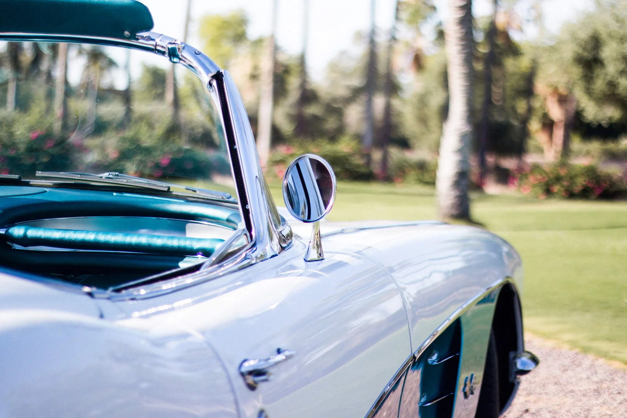 Close-up of a vintage white and turquoise 1959 Chevrolet Corvette convertible car with a side mirror and dashboard, set outdoors with trees and grass in the background.