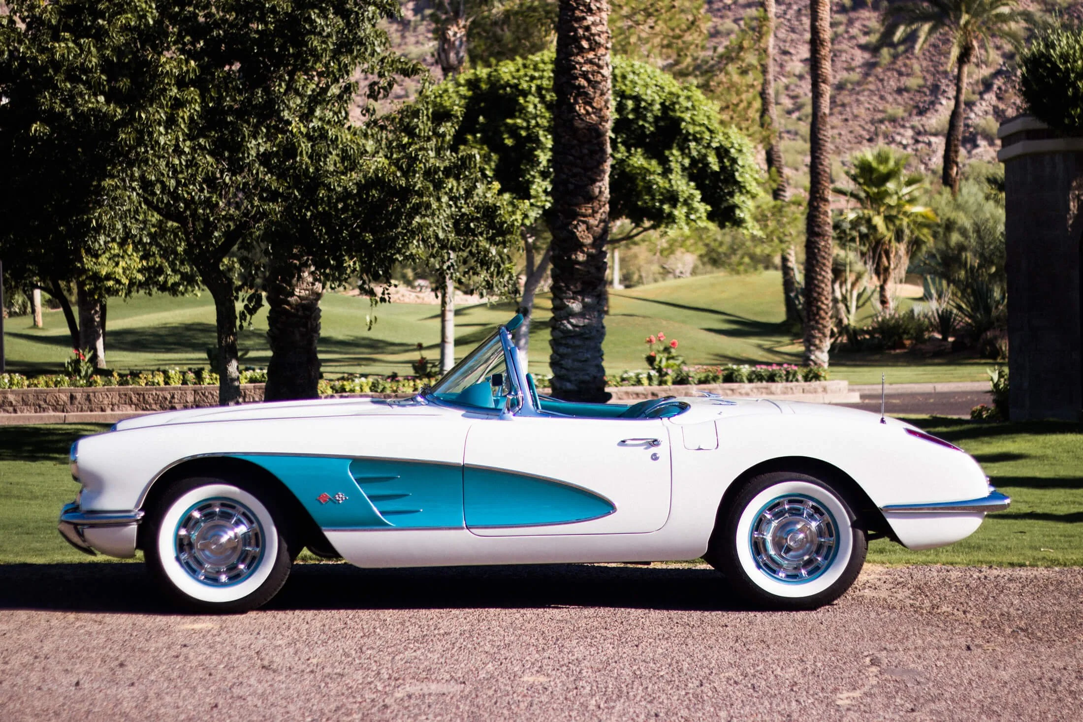 A classic white and turquoise 1959 Corvette convertible sports car parked on a gravel driveway with a lush green park, palm trees, and flowers in the background.