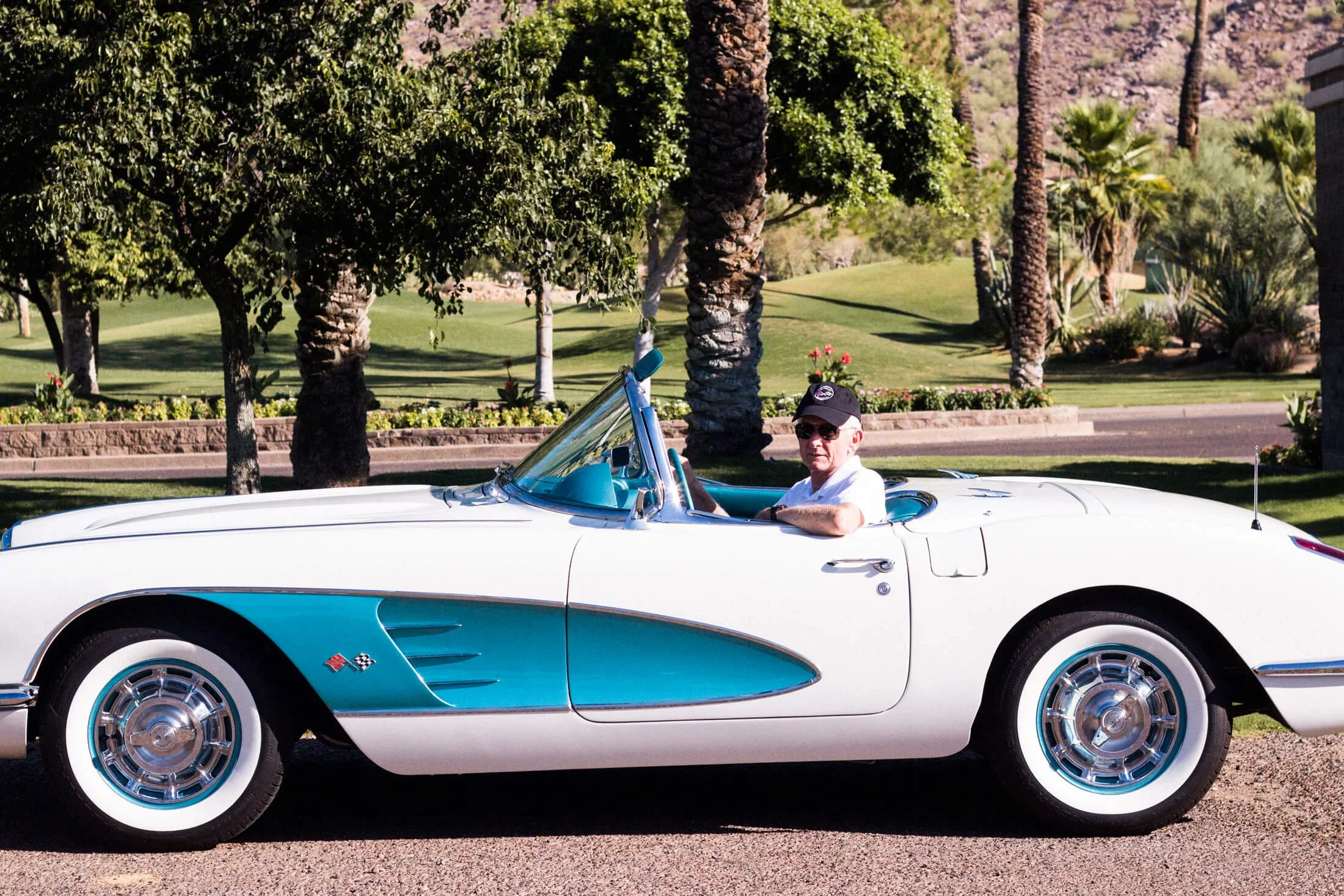 A man wearing sunglasses and a cap sitting in a vintage white and teal 1959 Chevrolet Corvette convertible car parked on a street with a park or garden in the background, featuring trees and grass.