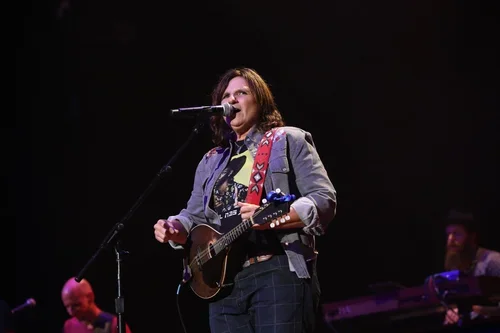 A woman performing on stage with a guitar, singing into a microphone, with musicians in the background.