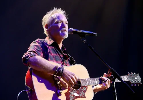 A man with gray hair and beard playing an acoustic guitar and singing into a microphone on stage