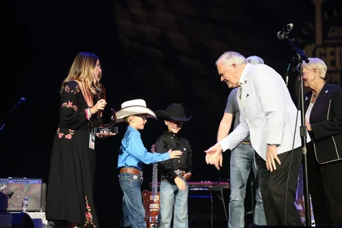 A man in a white blazer and two young boys, one wearing a cowboy hat, on stage receiving awards from a woman in a black dress with floral print at a ceremony.
