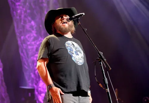 A man singing into a microphone on stage, wearing a black cowboy hat and a black t-shirt with a skull design.