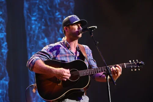 A man in a cap and plaid shirt playing an acoustic guitar and singing into a microphone on stage with blue lighting.