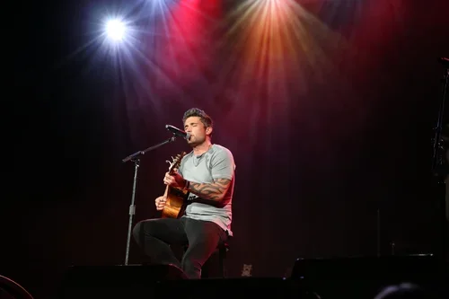 A man performing on stage with a guitar, sitting on a stool, in front of a microphone, with colorful stage lights overhead.