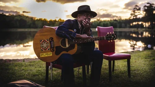 A man sitting outdoors by a lake at sunset, wearing a hat and dark clothing, playing an acoustic guitar with a 'Not Loud For' sticker, and covering his mouth with his hand.