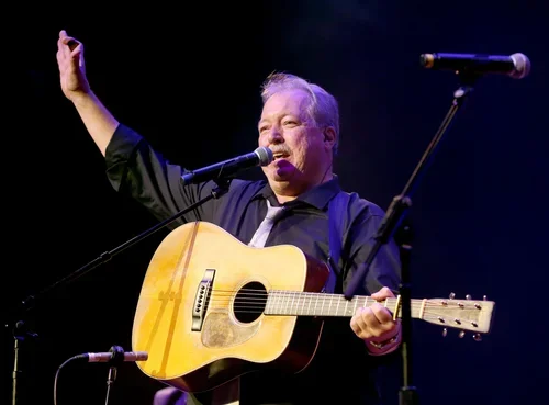 A man performing on stage with a guitar and a microphone, raising one arm.