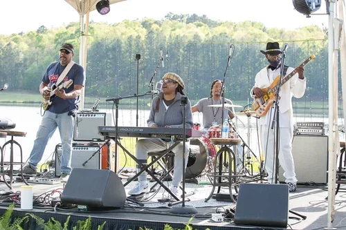 Band performing outdoors on a stage near a lake, with three musicians playing guitar, keyboard, and drums.