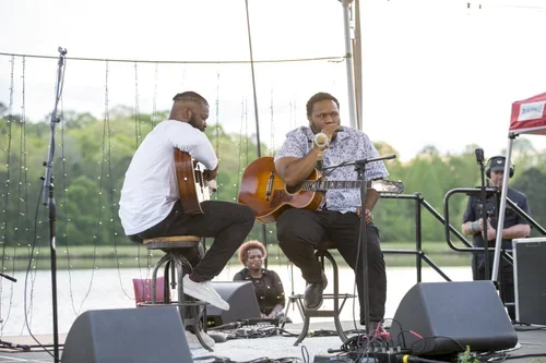 Two musicians playing guitars and singing on an outdoor stage near a body of water.