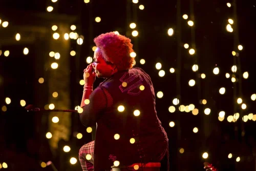 Person with pink curly hair sitting among hanging lights in a dark setting