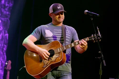 Man playing acoustic guitar on stage with microphone stand nearby.