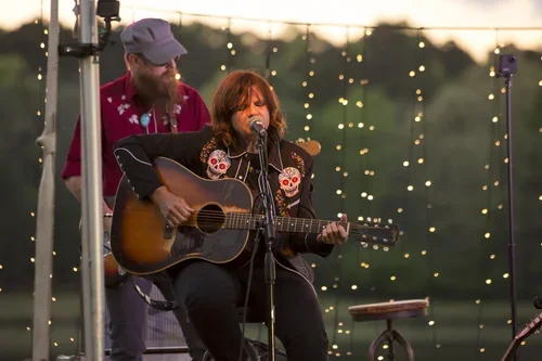 Woman playing guitar and singing into a microphone during an outdoor concert, with man standing behind her.