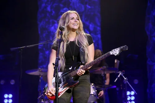 A woman with long blonde hair holding an electric guitar on stage with a drummer in the background, under blue stage lighting.