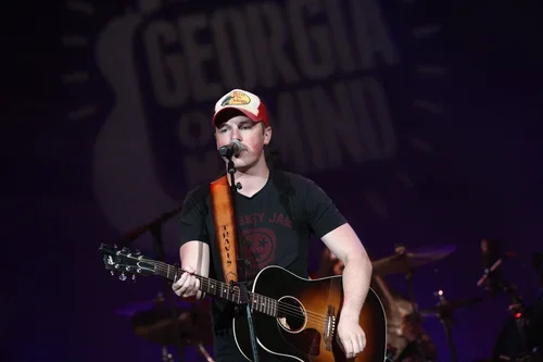 A young man performing on stage with a microphone, playing an acoustic guitar, wearing a red and white cap and a dark t-shirt with a graphic design.