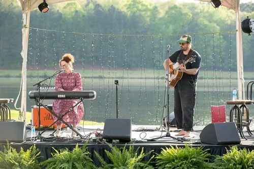 A man playing an acoustic guitar and a woman playing a keyboard performing outdoors on a stage near a body of water with a scenic green background.