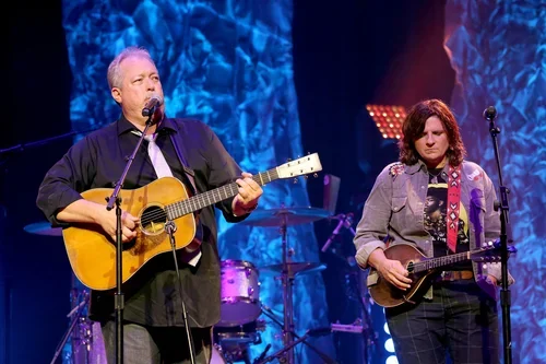 Two musicians performing on stage with guitars, a man singing into a microphone and a woman playing an electric guitar, with blue lighting and stage background.