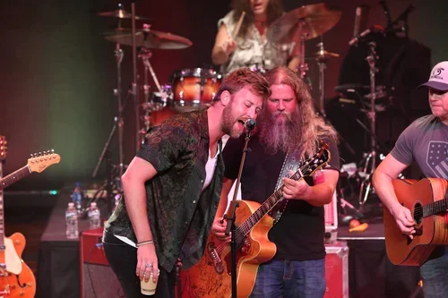 Musicians performing on stage, with two guitarists singing and playing guitars, and a drummer in the background.