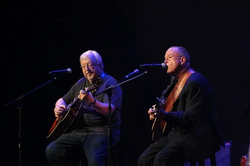 Two men playing acoustic guitars on a stage with microphones.