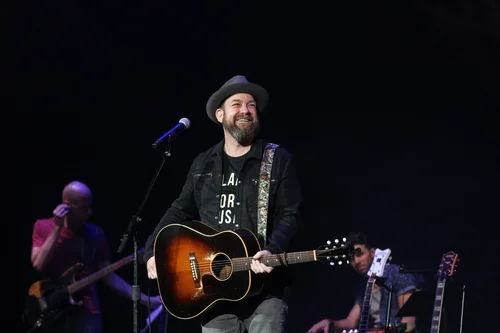 A man with a beard, wearing a hat and black jacket, is playing an acoustic guitar on stage, smiling. Two other musicians are in the background, one with a bass guitar, the other with an electric guitar, performing under stage lights.