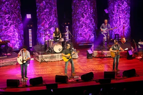 A band performing live on stage with three guitarists, a drummer, and a keyboard player. The stage has purple lighting and textured purple backdrops.